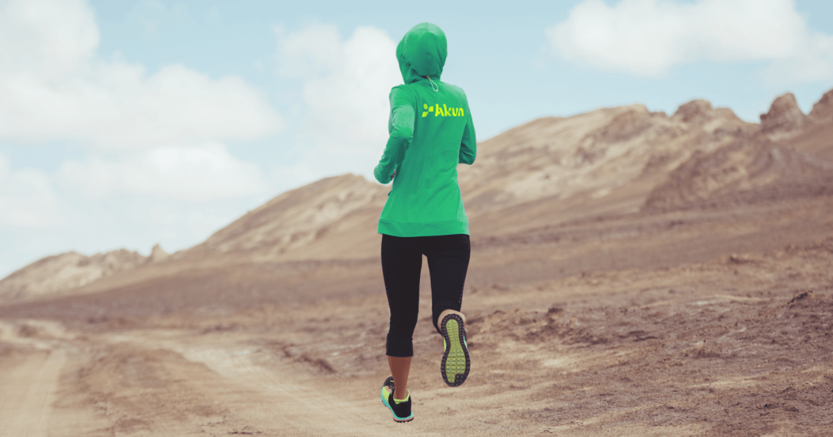 A woman wearing a green hoodie with the Akun logo runs through a scenic desert landscape.